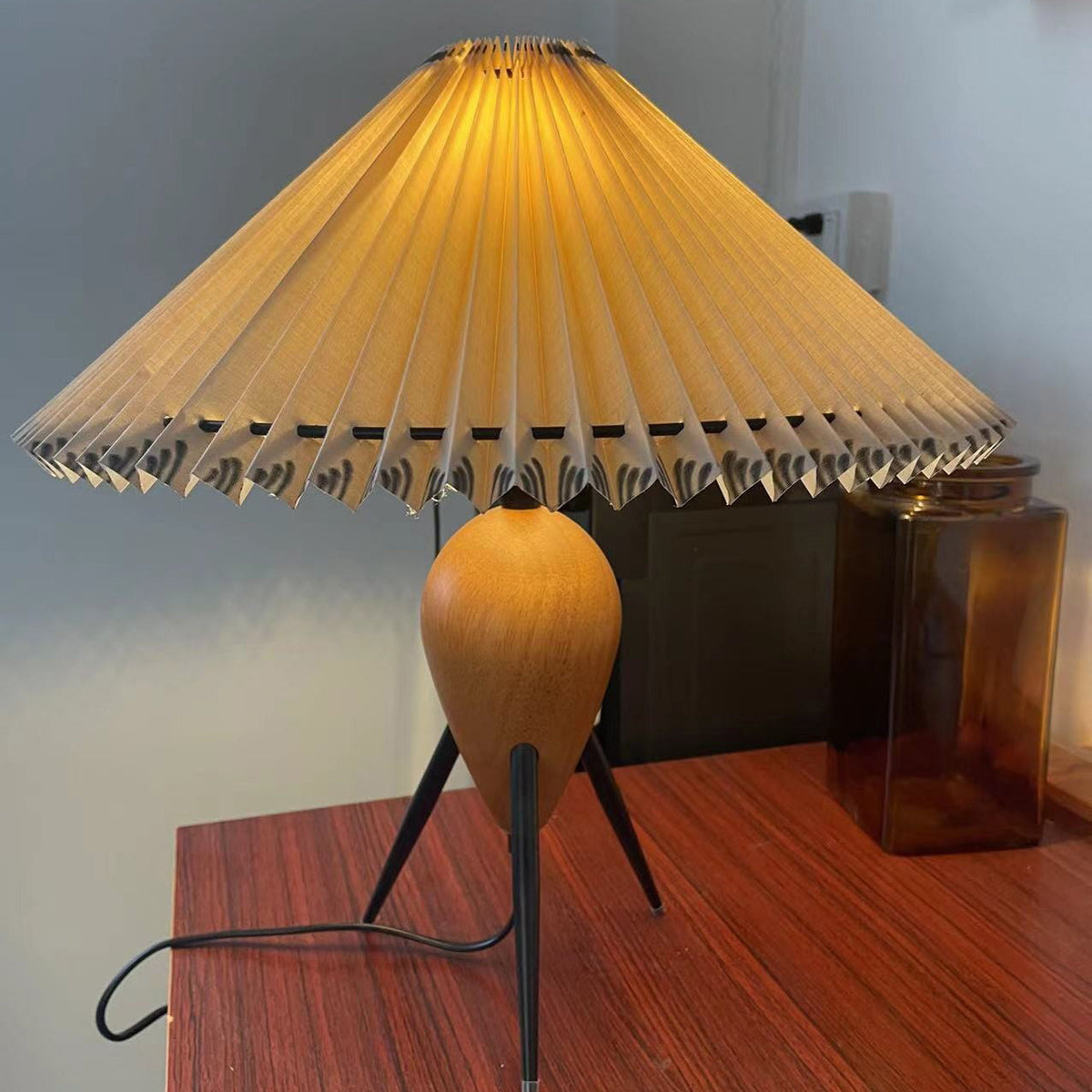 Looking up at the wide pleated shade of this Clérachic table lamp, lit, on a red-brown wood table beside a glass jar.