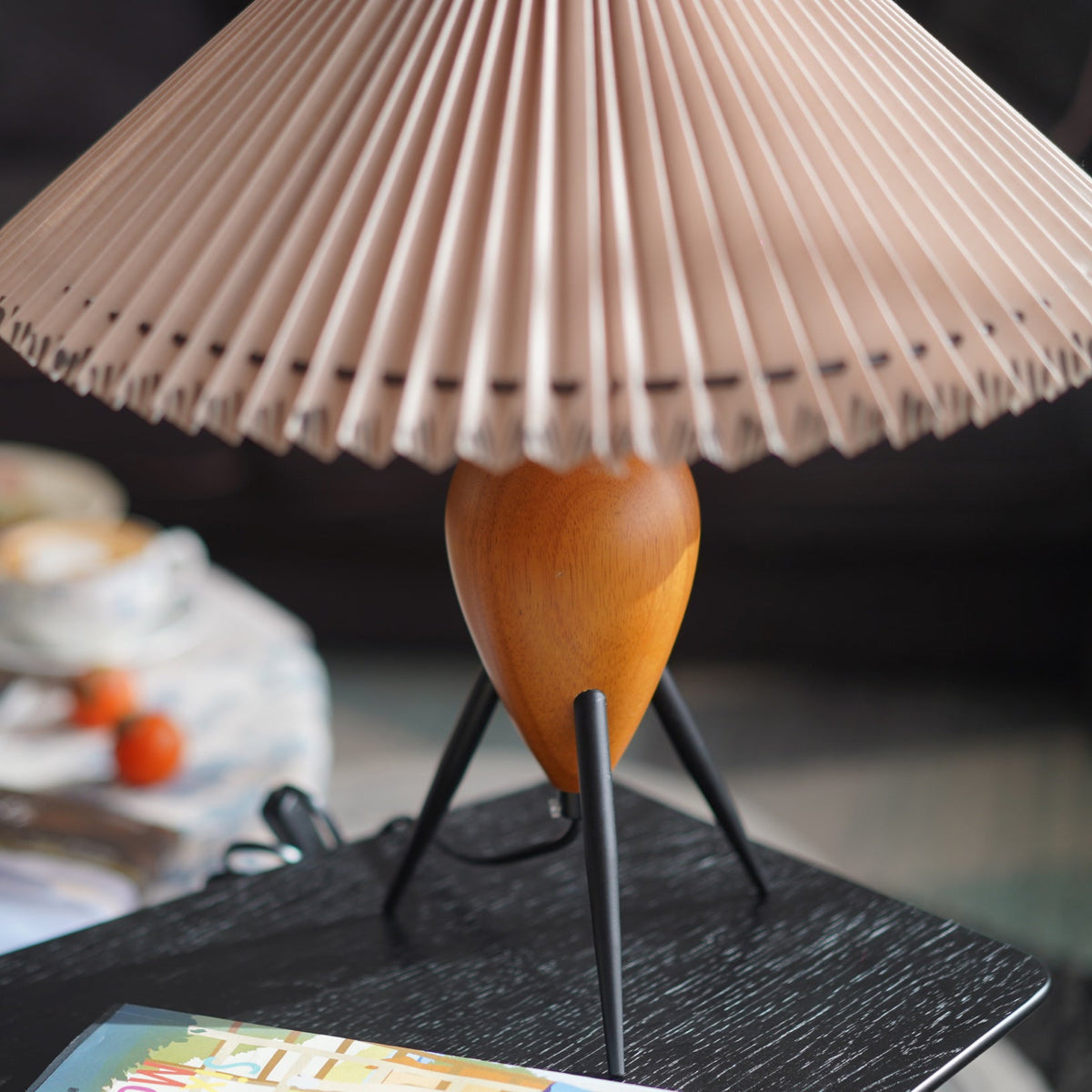 Tight view of a Clérachic table lamp's pleated shade and wood base, lit, with a book and a dinner table behind.