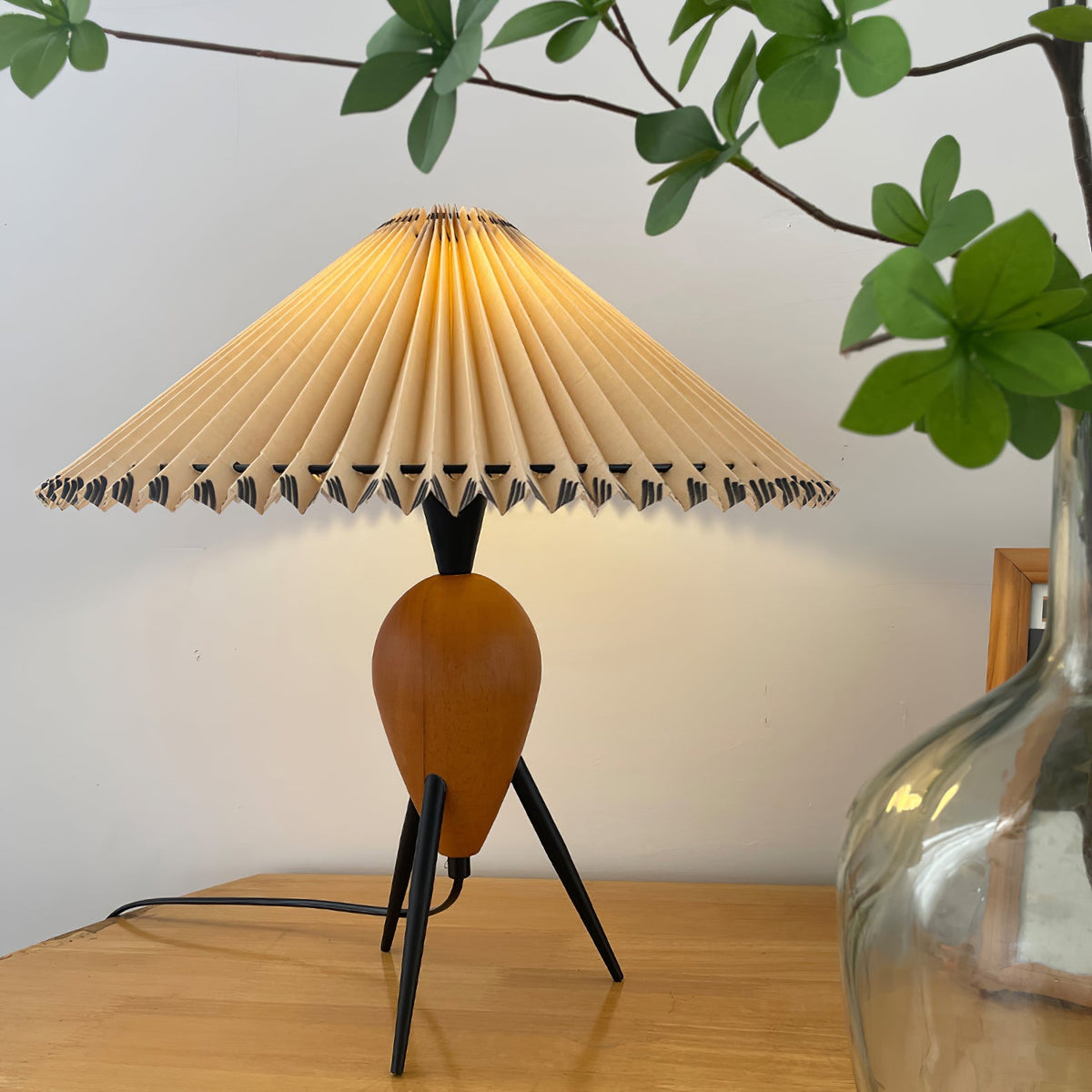 This Clérachic table lamp sits low on a honey wood table, a green branch and a glass jug blurred around its pleated shade.