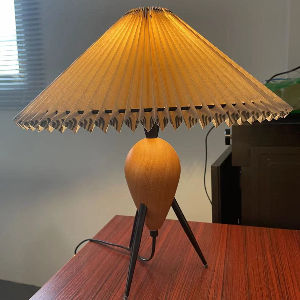 This Clérachic table lamp glows over a red-brown wood table, its legs splayed and a black cord trailing to the left.