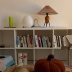 On a shelf above a unit of books, this Clérachic table lamp stands in a living room beside a white paper lantern.