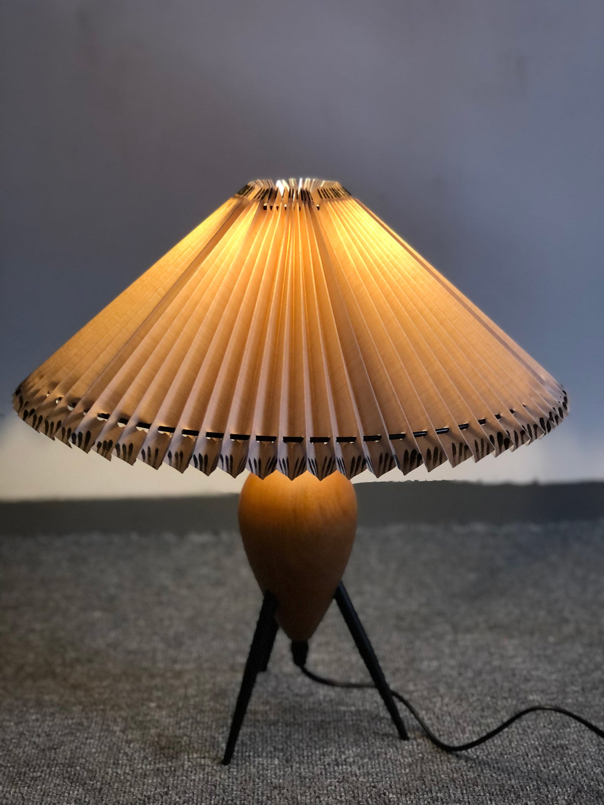 Lit and dark-toned, a Clérachic table lamp stands on a grey floor against a blue-grey backdrop.
