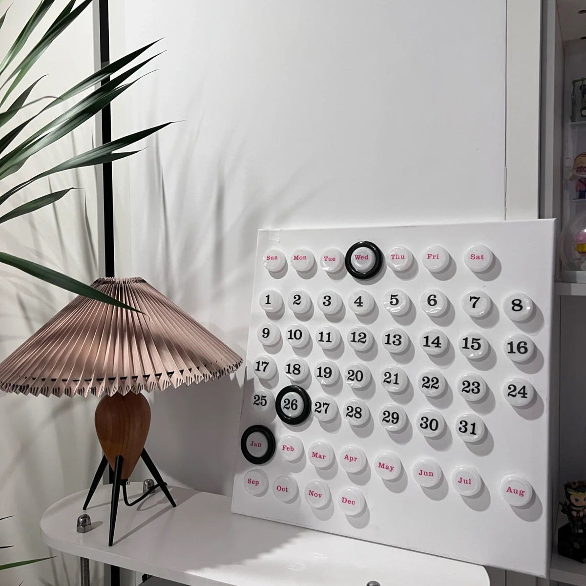 A Clérachic table lamp beside a white wall calendar with round tiles, a leafy plant leaning in from the left.