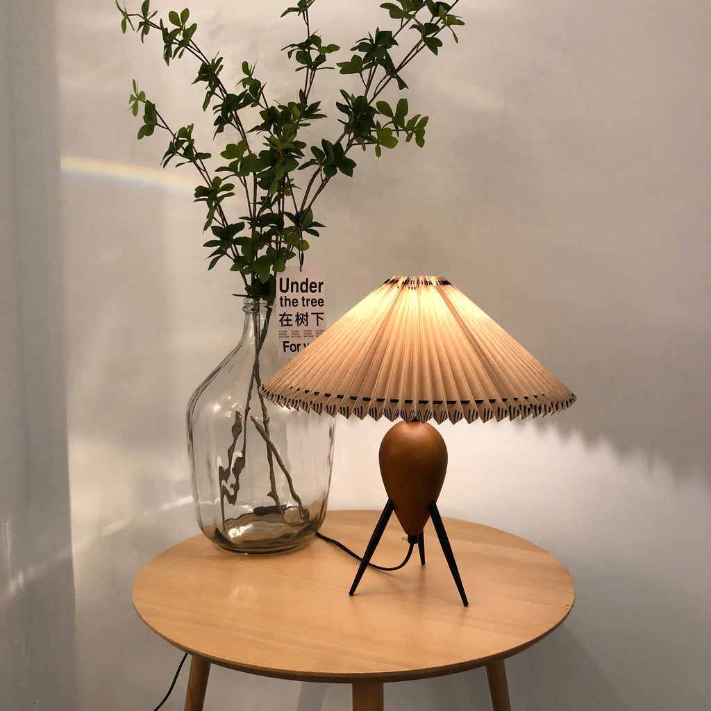 Lit Clérachic table lamp on a round wood table, a glass jar of leafy branches and a small card behind it.