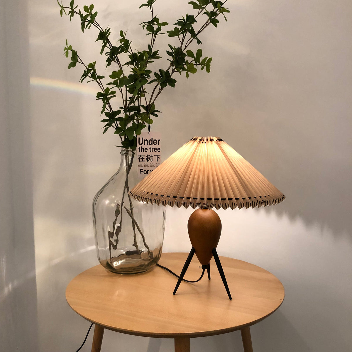 Lit Clérachic table lamp on a round wood table, a glass jar of leafy branches and a small card behind it.
