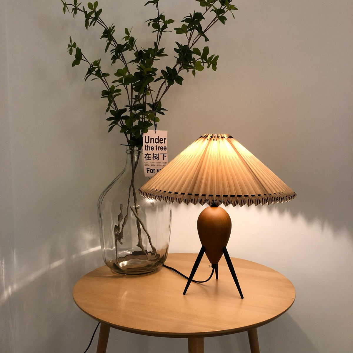 A lit Clérachic table lamp on a round wood table, warm light on the pleats, with a tall jar of green branches at its side.