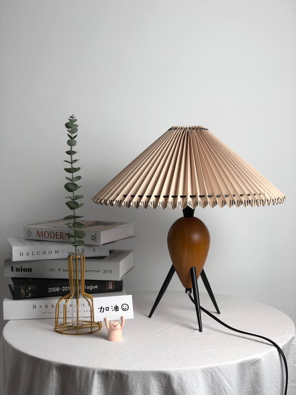 This Clérachic table lamp stands on a white draped table with a gold wire vase, eucalyptus and stacked books, shade unlit.