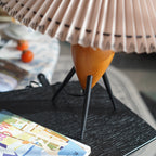 Close view of a Clérachic table lamp's wood base and black legs beneath the pleated shade, on a black tray by a book.