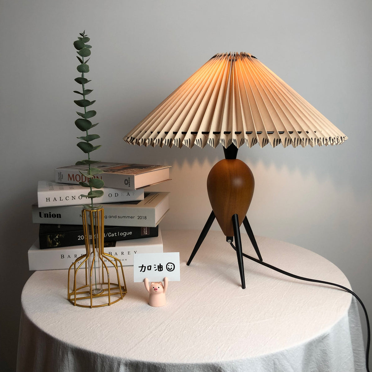 Warm-lit Clérachic table lamp on a draped round table, a green eucalyptus stem and books at its left.