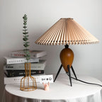 This Clérachic table lamp on a white-draped table, with a stack of books, a wire vase and a small pink figure in a bright room.