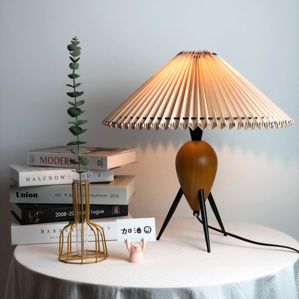 A glowing Clérachic table lamp next to a stack of design books, a green stem and a gold wire vase.