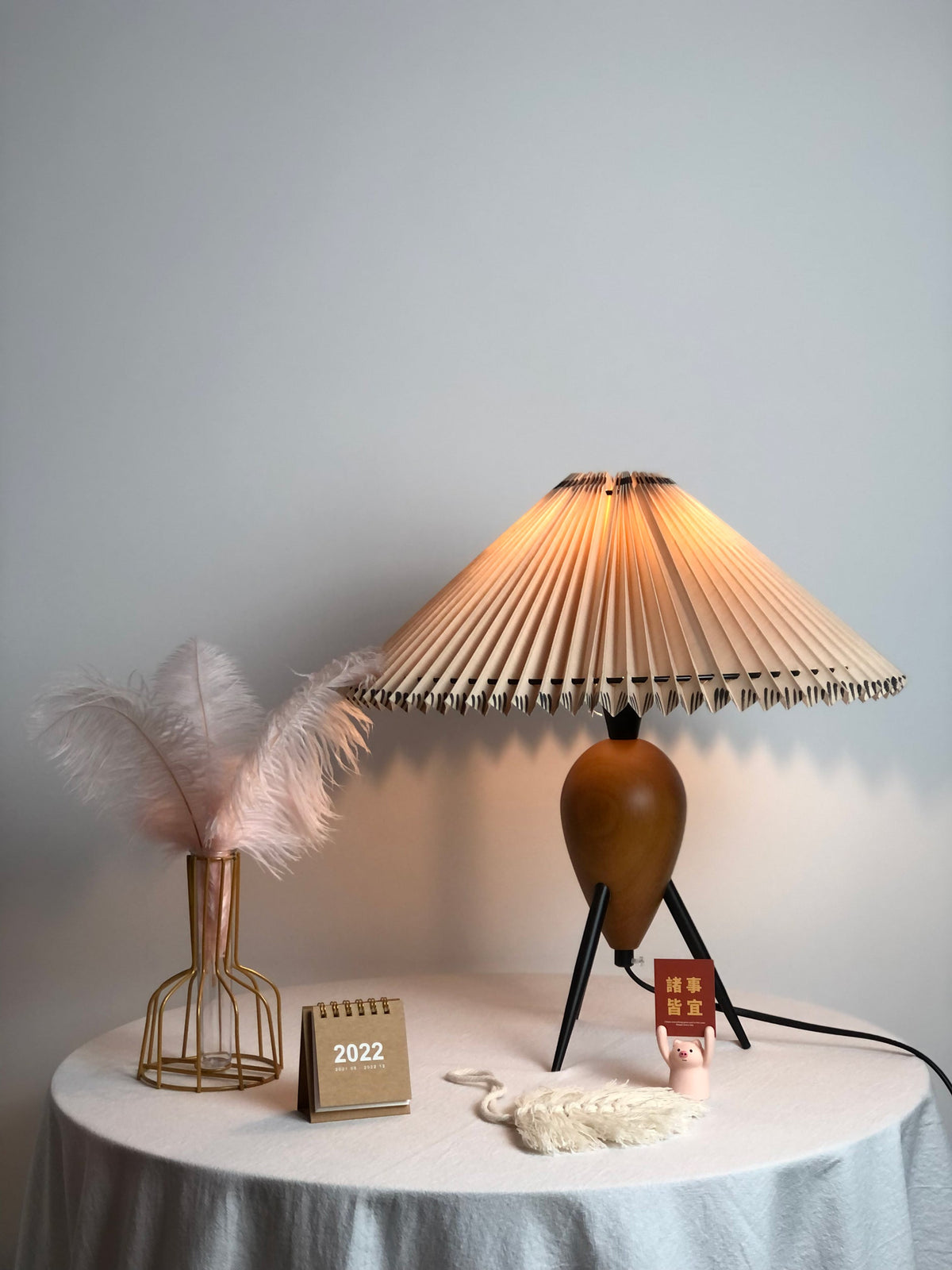 A pink feather in a gold wire vase, a mini calendar and a woven tassel sit beside this lit Clérachic table lamp.