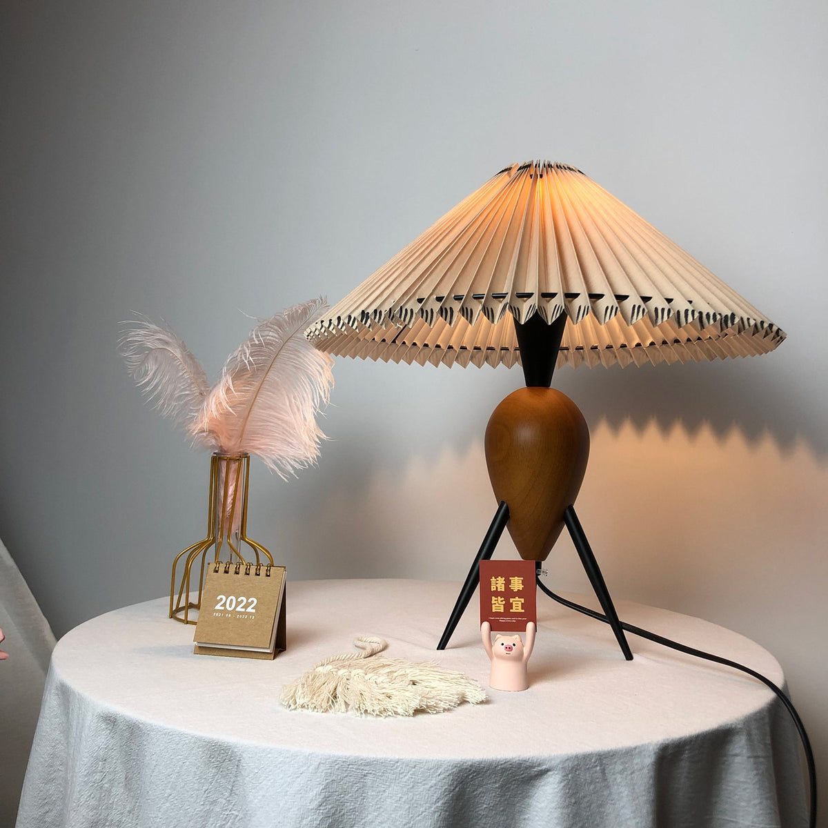 This Clérachic table lamp on a draped table, lit, with the pink feather vase and mini calendar placed close to the base.