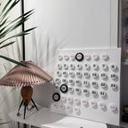 A Clérachic table lamp by a white calendar board and tall plant, with its pleated shade in a soft blush tone.