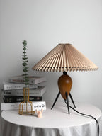 Beside books, a gold wire vase and a green stem, this Clérachic table lamp stands on a draped table, shade unlit.