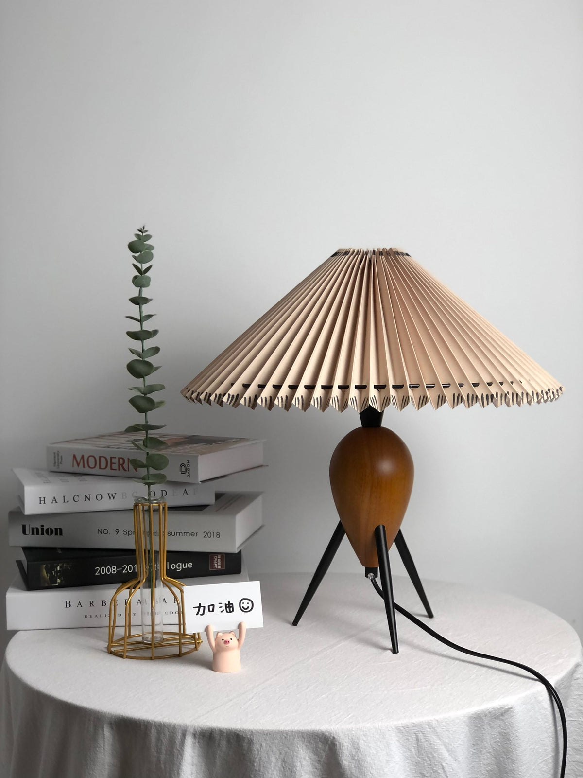 Beside books, a gold wire vase and a green stem, this Clérachic table lamp stands on a draped table, shade unlit.