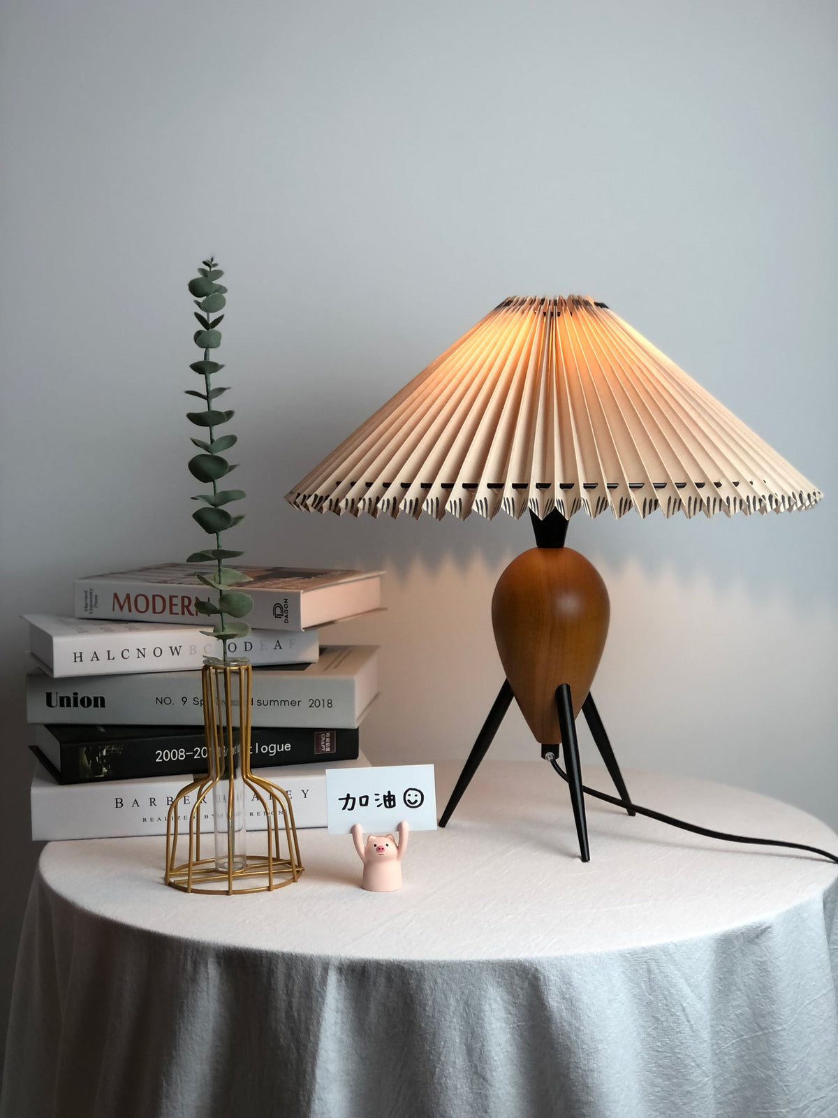 A bright Clérachic table lamp on a table with a pale grey drape, a stem of eucalyptus and books on its left.