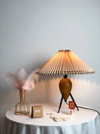This Clérachic table lamp, lit, with a pink feather in a gold wire vase and a mini calendar on a round white table.