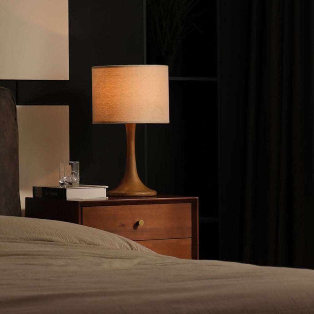 Lit Clérachic table lamp glowing amber on a wooden bedside chest, with a glass of water and a book beside a made bed.