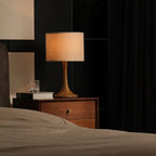 Lit Clérachic table lamp glowing amber on a wooden bedside chest, with a glass of water and a book beside a made bed.