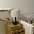 A black-bodied Clérachic table lamp with a beige drum shade, on a pale wooden bedside unit beside a cream sofa.