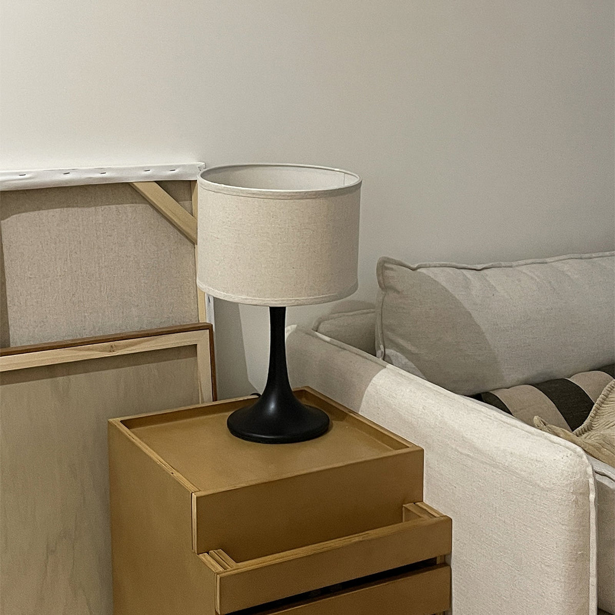 A black-bodied Clérachic table lamp with a beige drum shade, on a pale wooden bedside unit beside a cream sofa.