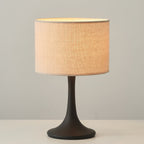 Lit Clérachic table lamp in black, its beige drum shade glowing peach on a white tabletop against a grey wall.