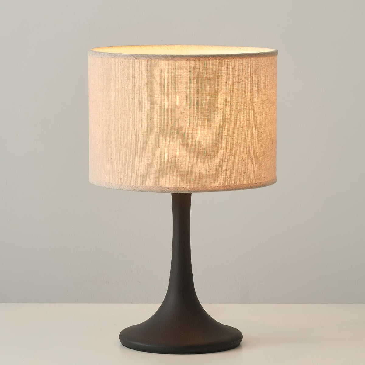 Lit Clérachic table lamp in black, its beige drum shade glowing peach on a white tabletop against a grey wall.