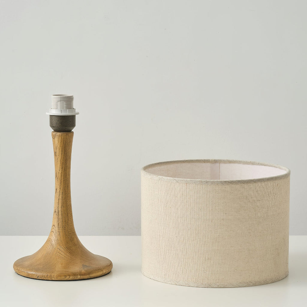 The wood-coloured base of this Clérachic table lamp, with its bulb socket showing, stands next to the beige linen drum shade.