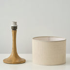 The wood-coloured base of this Clérachic table lamp, with its bulb socket showing, stands next to the beige linen drum shade.