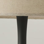 Close view of the black turned column of this Clérachic table lamp meeting the underside of its woven beige drum shade.