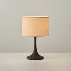 Clérachic table lamp in black, lit on a white surface, with its beige drum shade glowing above a slim, flared black stem.