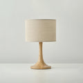 Front view of this Clérachic table lamp in a wood colour, with a beige woven drum shade above a flared turned-wood foot.