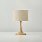 Front view of this Clérachic table lamp in a wood colour, with a beige woven drum shade above a flared turned-wood foot.
