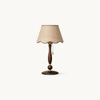 Table lamp with a scalloped beige woven shade edged in brown, a turned walnut stem and a round dark base, on a white backdrop; a Clérachic lamp.