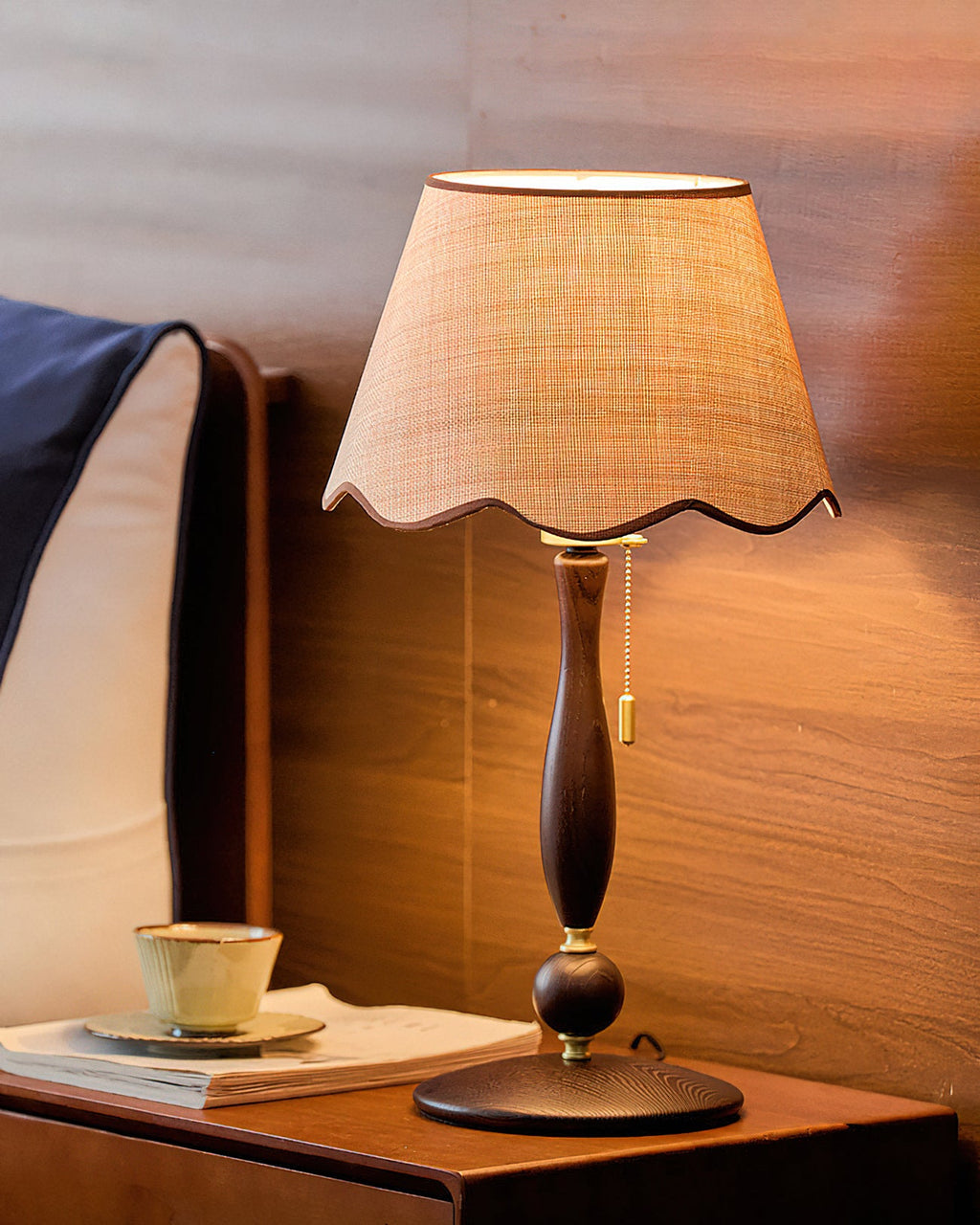Lit scalloped shade glowing above a turned wooden stem, beside a teacup and a magazine on a walnut bedside table; from Clérachic.