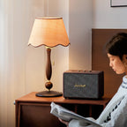 Warm light from the shade falls on a walnut bedside table with a speaker while a person reads in the corner; a Clérachic table lamp.