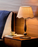 Lit lamp on a walnut bedside table with a teacup, in front of a dark headboard and a navy pillow; a Clérachic design.