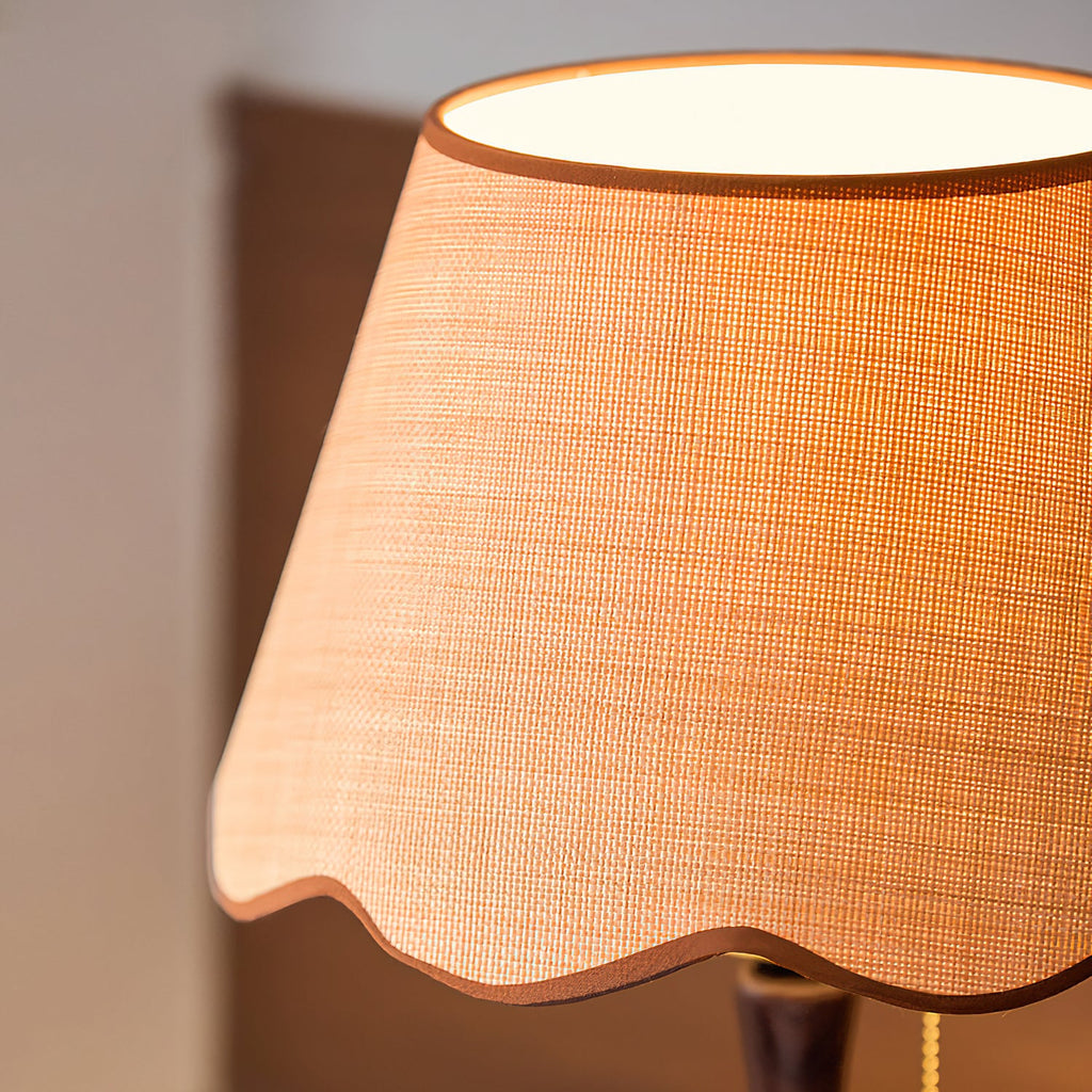 Close view of the woven beige shade, with its scalloped brown trim and a glow at the top opening; a Clérachic table lamp.