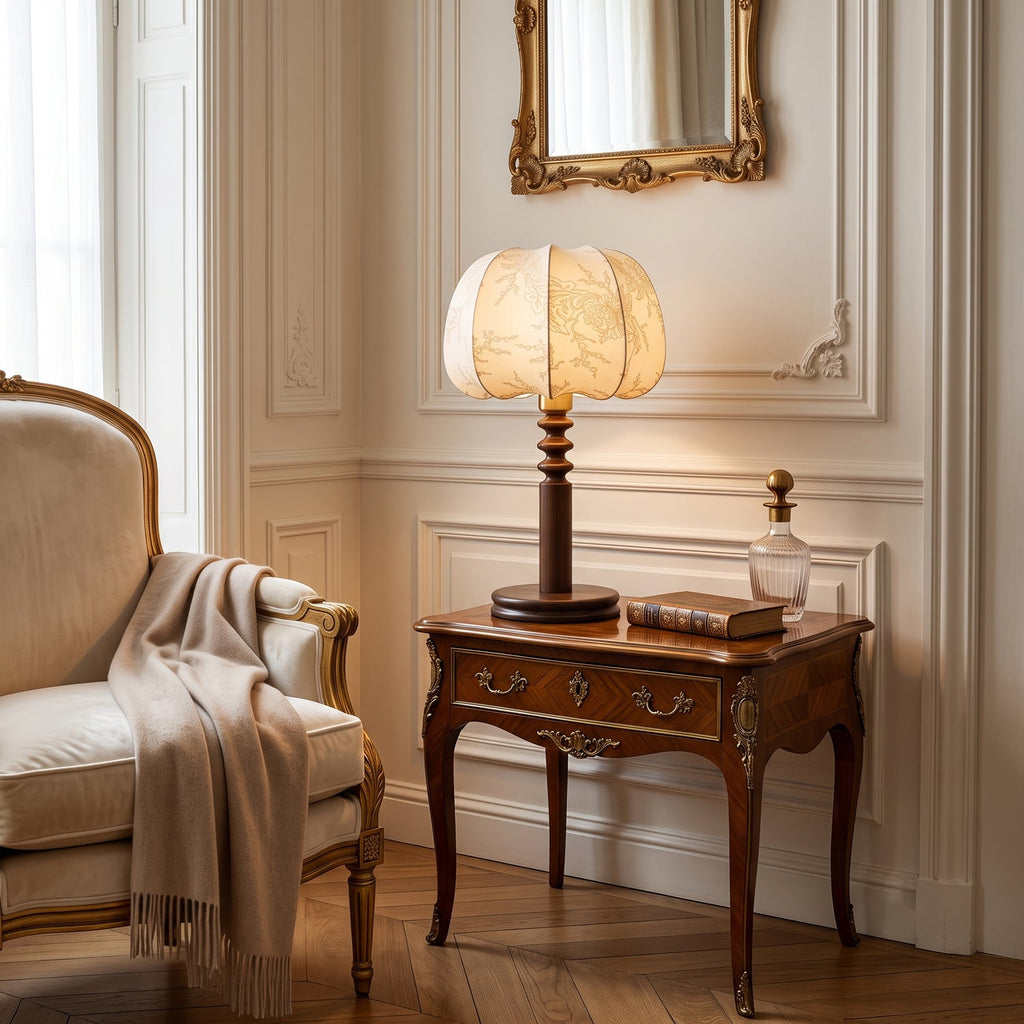 Lit Clérachic table lamp on a carved dark-wood side table beside a cream armchair with a throw, under a gilt mirror.