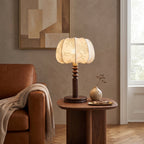 Warm light from a Clérachic table lamp on a round walnut side table, beside a tan leather armchair and a small bud vase.