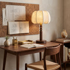 A Clérachic table lamp lit on a walnut writing desk, with pinned drawings on a cork board, a brass tray and a woven-seat chair.