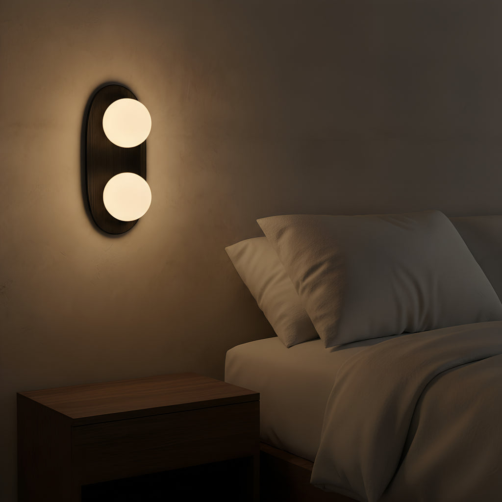 The black two-light wall sconce mounted at head height beside a bed at night, both globes glowing warm