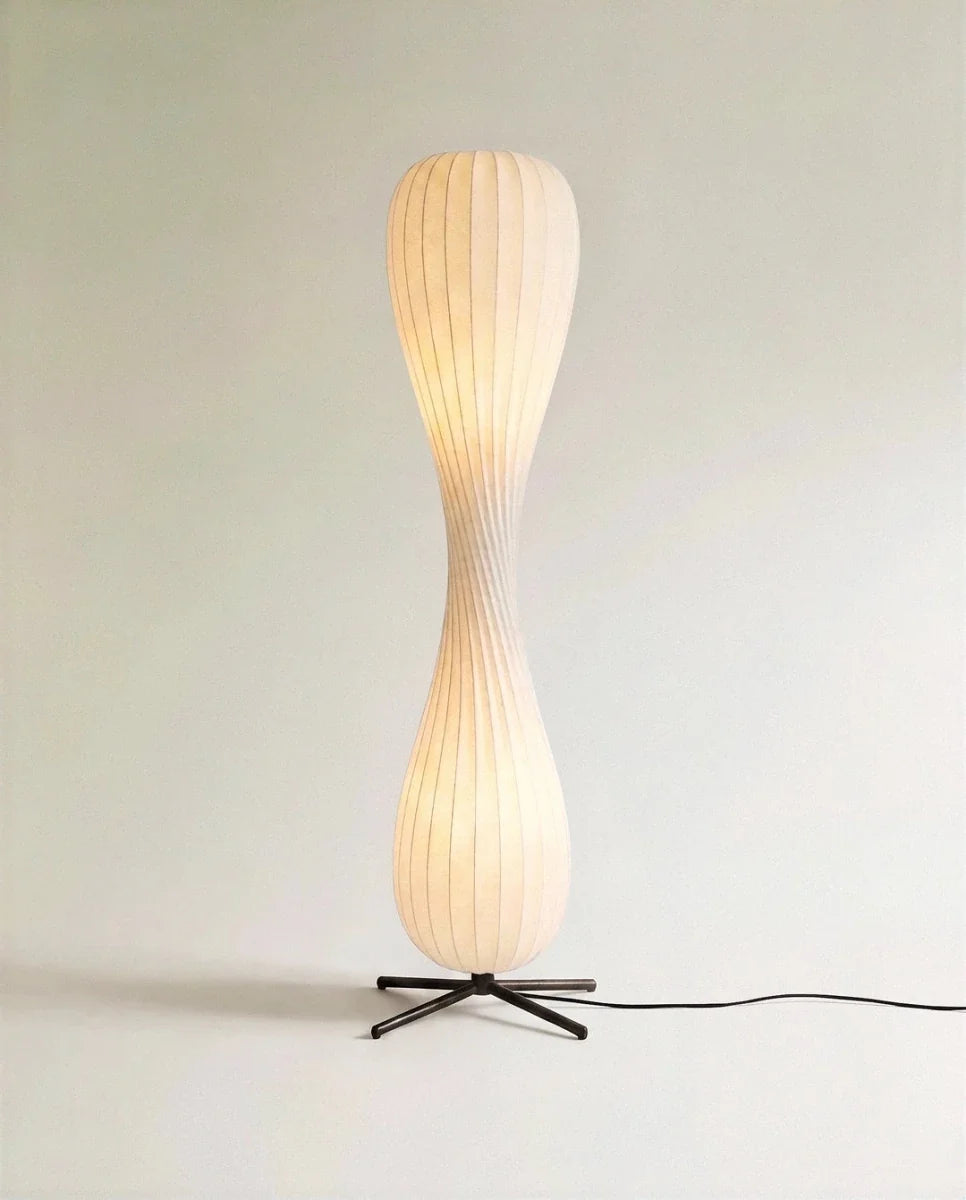 Clérachic twisted hourglass floor lamp glowing warm from within its ribbed shade, black cable trailing from the cross base.