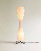 Clérachic twisted hourglass floor lamp glowing warm from within its ribbed shade, black cable trailing from the cross base.