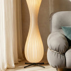Clérachic floor lamp lit in a sunny corner, its ribbed shade between linen curtains and a grey bouclé sofa arm.
