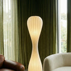 Clérachic tall ribbed floor lamp glowing in front of green velvet curtains, beside a cream bouclé sofa.