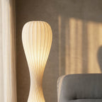 Clérachic ribbed floor lamp glowing in late sun beside a grey armchair, against a plaster wall with soft shadows.