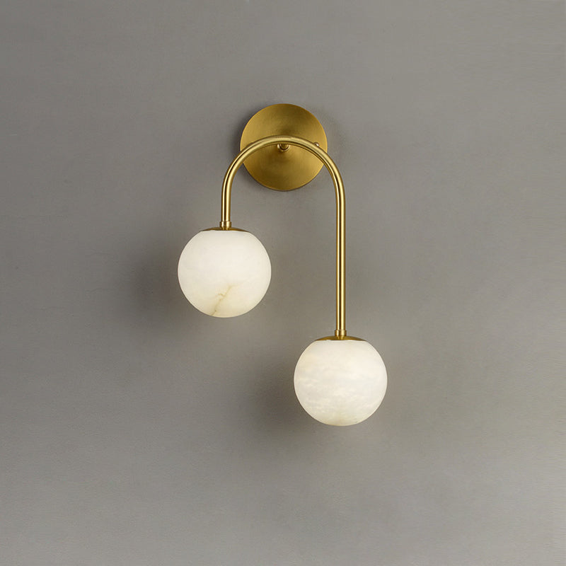 Clérachic Two-Globe Alabaster Wall Sconce in brass on a plain grey wall, its high and low stone globes staggered along one curved arm.