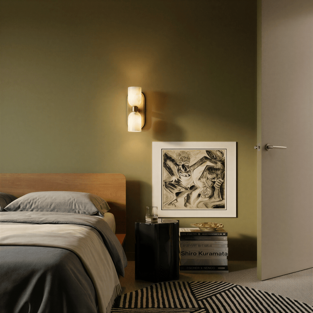 Clérachic dual-shade wall sconce lit on an olive bedroom wall above a black nightstand, beside a framed print and a wood headboard.